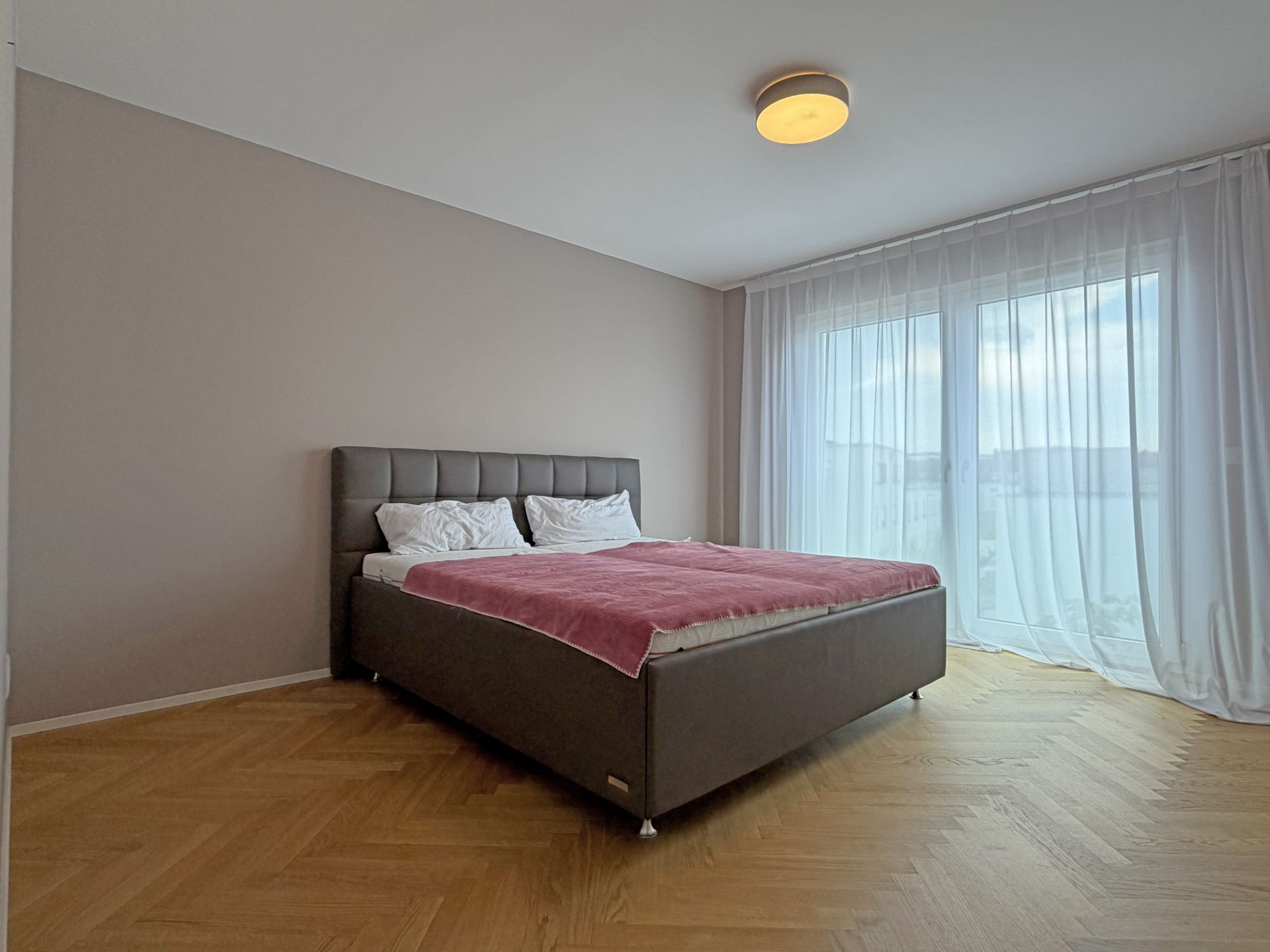 Helles Schlafzimmer mit Doppelbett, rosa Decke und großen Fenstern mit weißen Vorhängen.