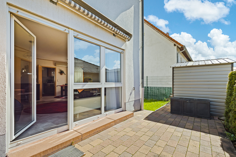 Terrasse mit geöffnetem Glas-Schiebetür zur Wohnung, gepflasterter Boden, kleines Gartenhaus rechts.