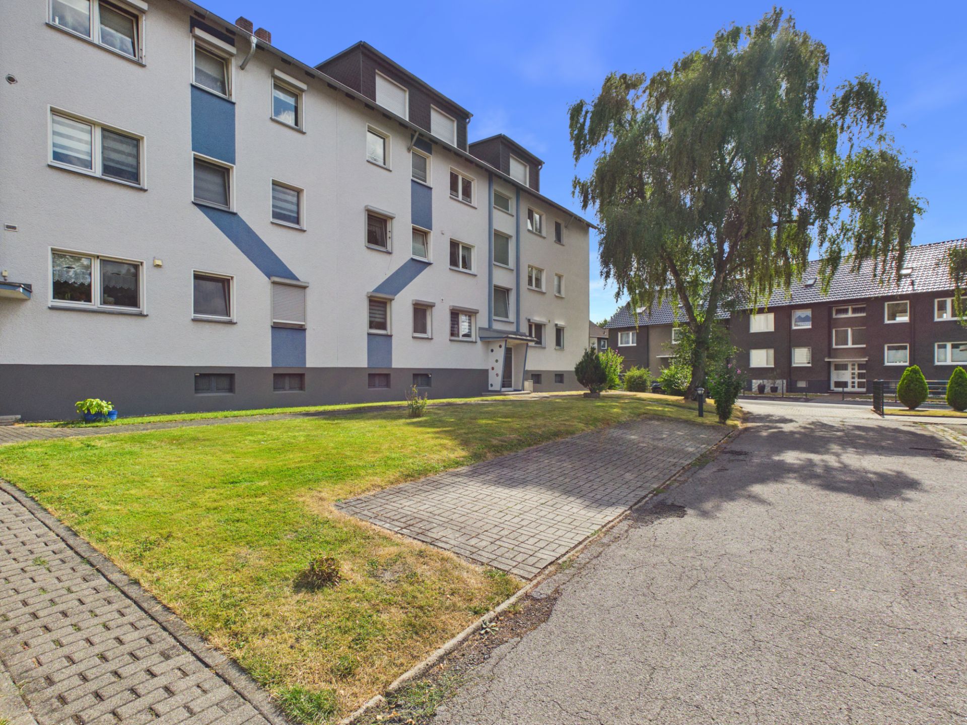 Mehrfamilienhaus mit Rasenfläche, Gehweg und Baum im Vordergrund.