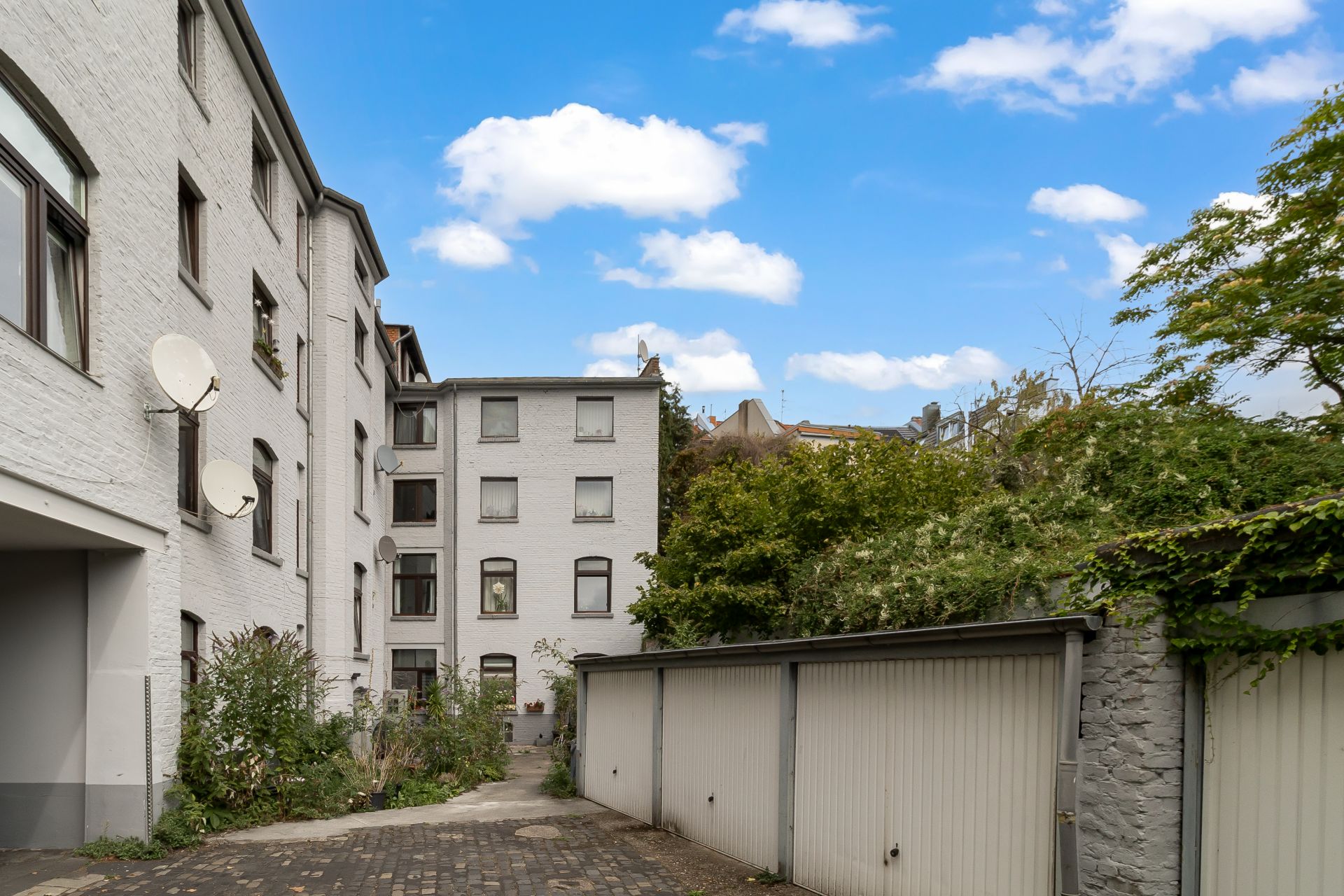 Innenhof mit weißen Wohnhäusern, Garagen und grünen Büschen unter blauem Himmel.