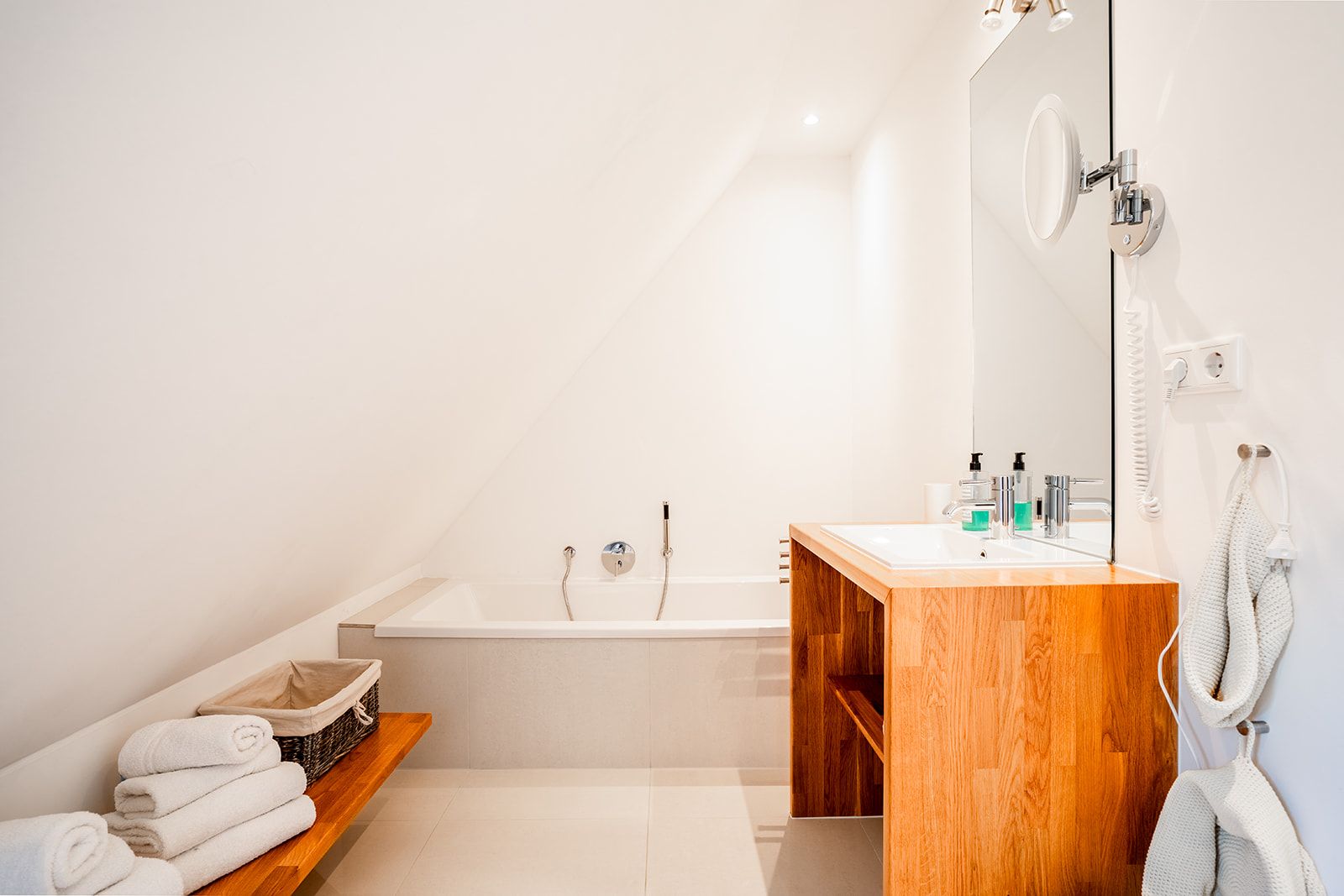 Helles Bad mit Dachschräge, Badewanne, Holzwaschtisch und einem Regal mit Handtüchern.