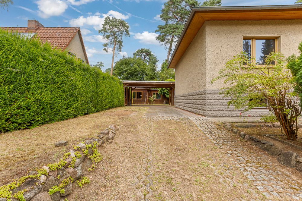 Gepflasterte Einfahrt neben einem Haus mit Garten, Hecke und Carport im Hintergrund.