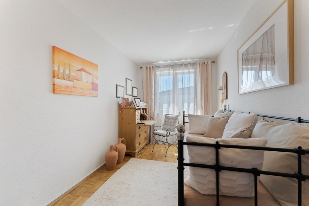 Helles Wohnzimmer mit Sofa, Holzmöbeln, Bildern an den Wänden und großen Fenstern mit Vorhängen.