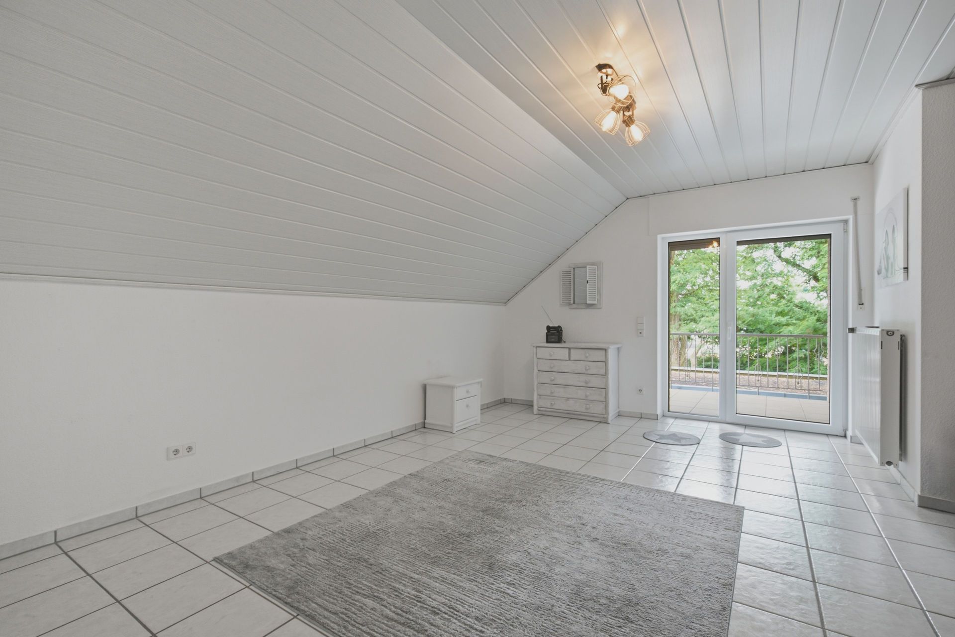 Helles Zimmer mit Schräge, Fliesenboden, Teppich, Kommode und Zugang zum Balkon.