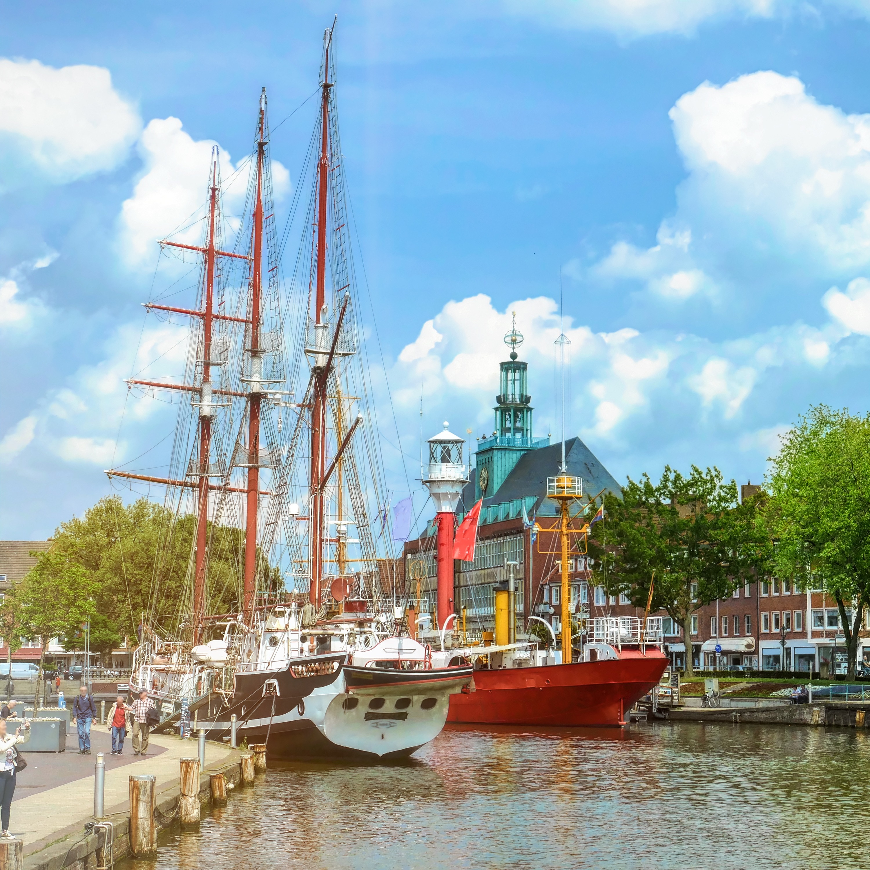 Zwei Segelschiffe und ein rotes Schiff liegen an einem Hafen, im Hintergrund Gebäude und ein Kirchturm.