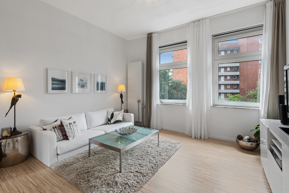 Helles Wohnzimmer mit weißem Sofa, Glastisch, Teppich und großen Fenstern.