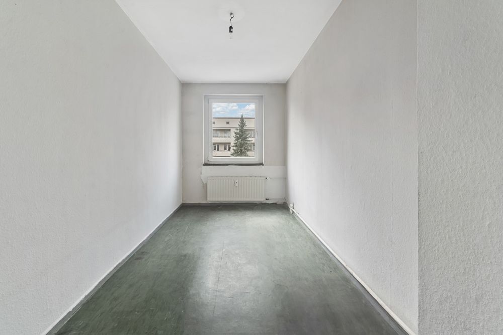 Leerer, schmaler Raum mit Fenster, Heizung und grünem Bodenbelag.