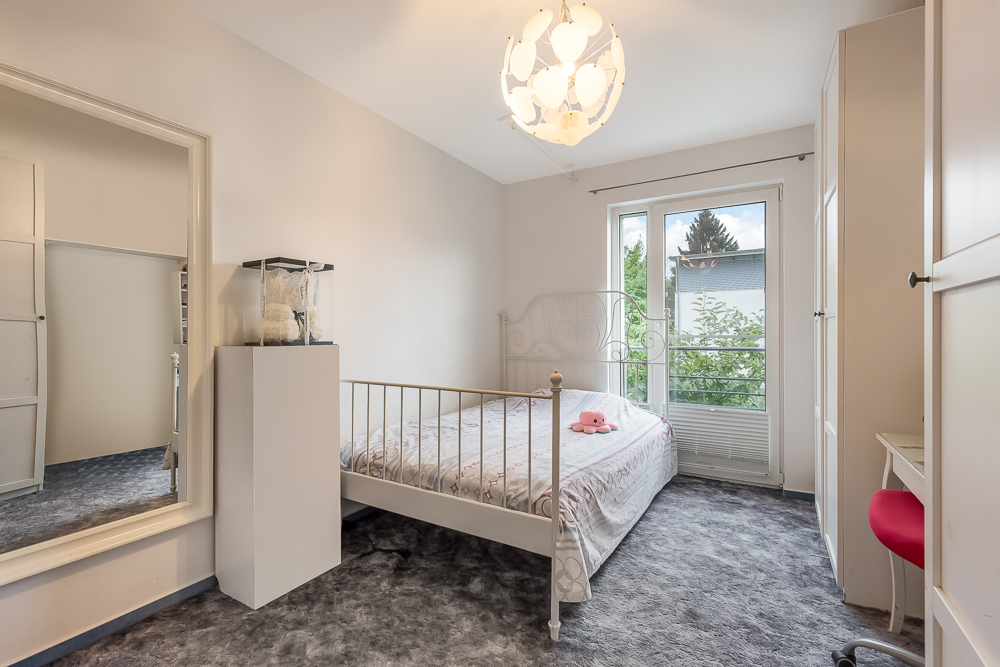 Helles Schlafzimmer mit großem Fenster, Bett, Kleiderschrank, Spiegel und Schreibtisch mit rotem Stuhl.