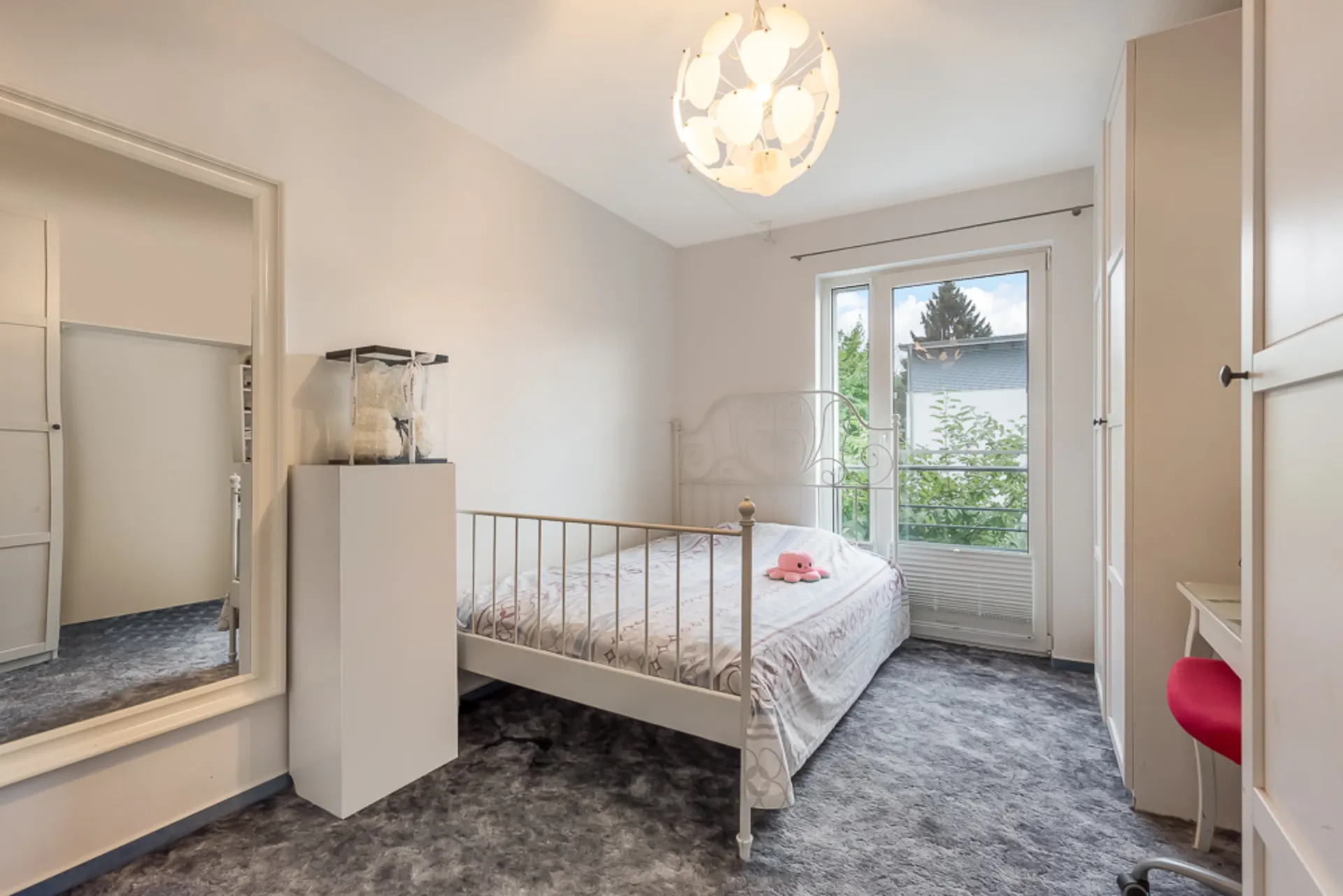 Helles Schlafzimmer mit großem Fenster, Bett, Kleiderschrank, Spiegel und Schreibtisch mit rotem Stuhl.