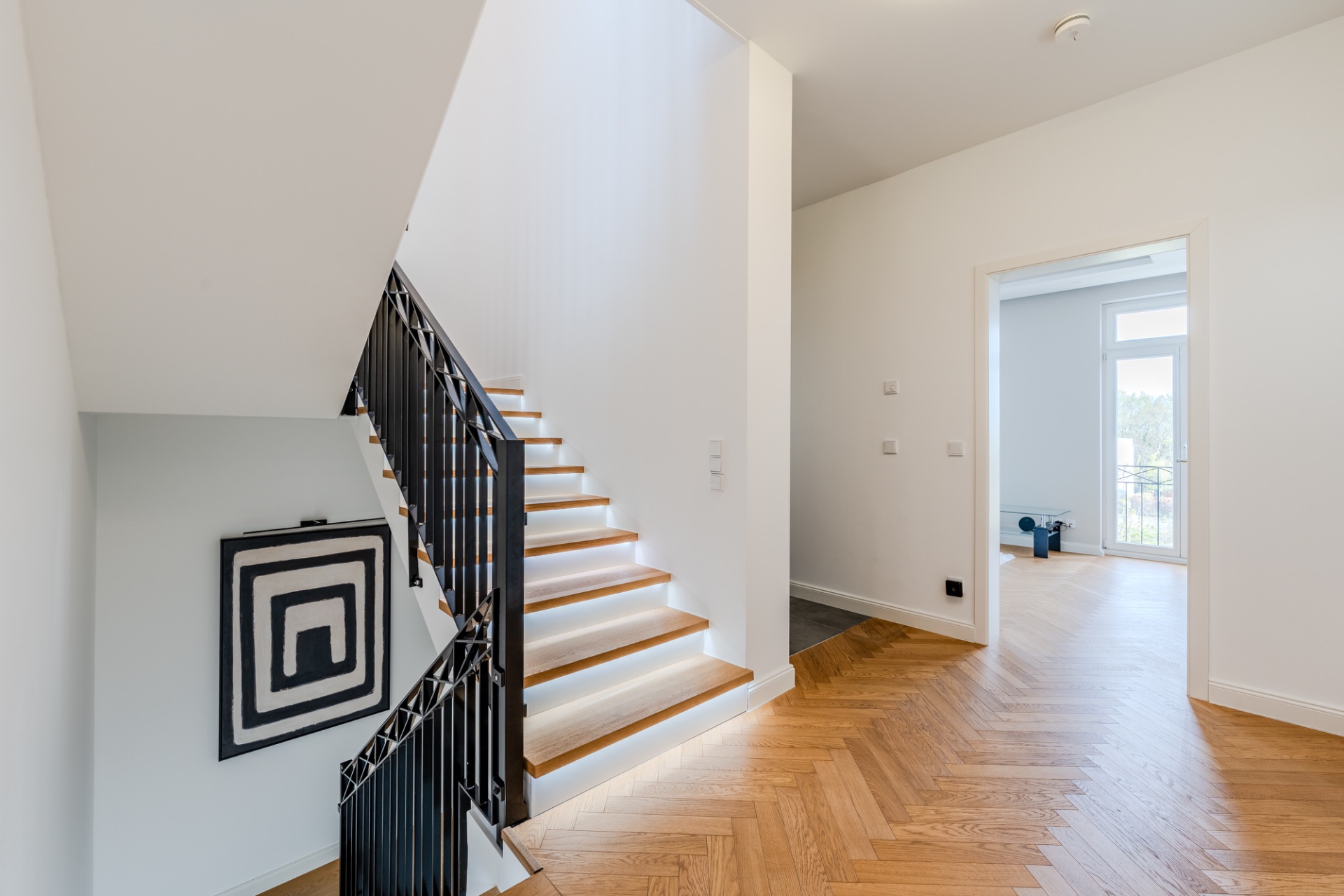 Heller Flur mit Holztreppe, Parkettboden und abstraktem Bild an der Wand.