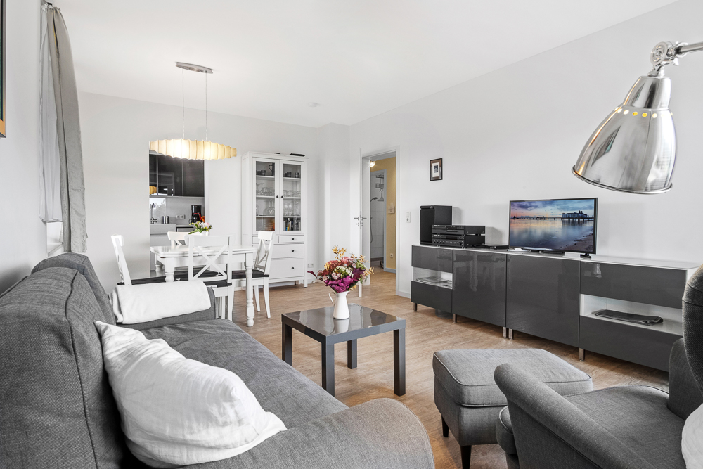 Modernes Wohnzimmer mit grauem Sofa, Sessel, Couchtisch, TV, Esstisch und Küchenblick.