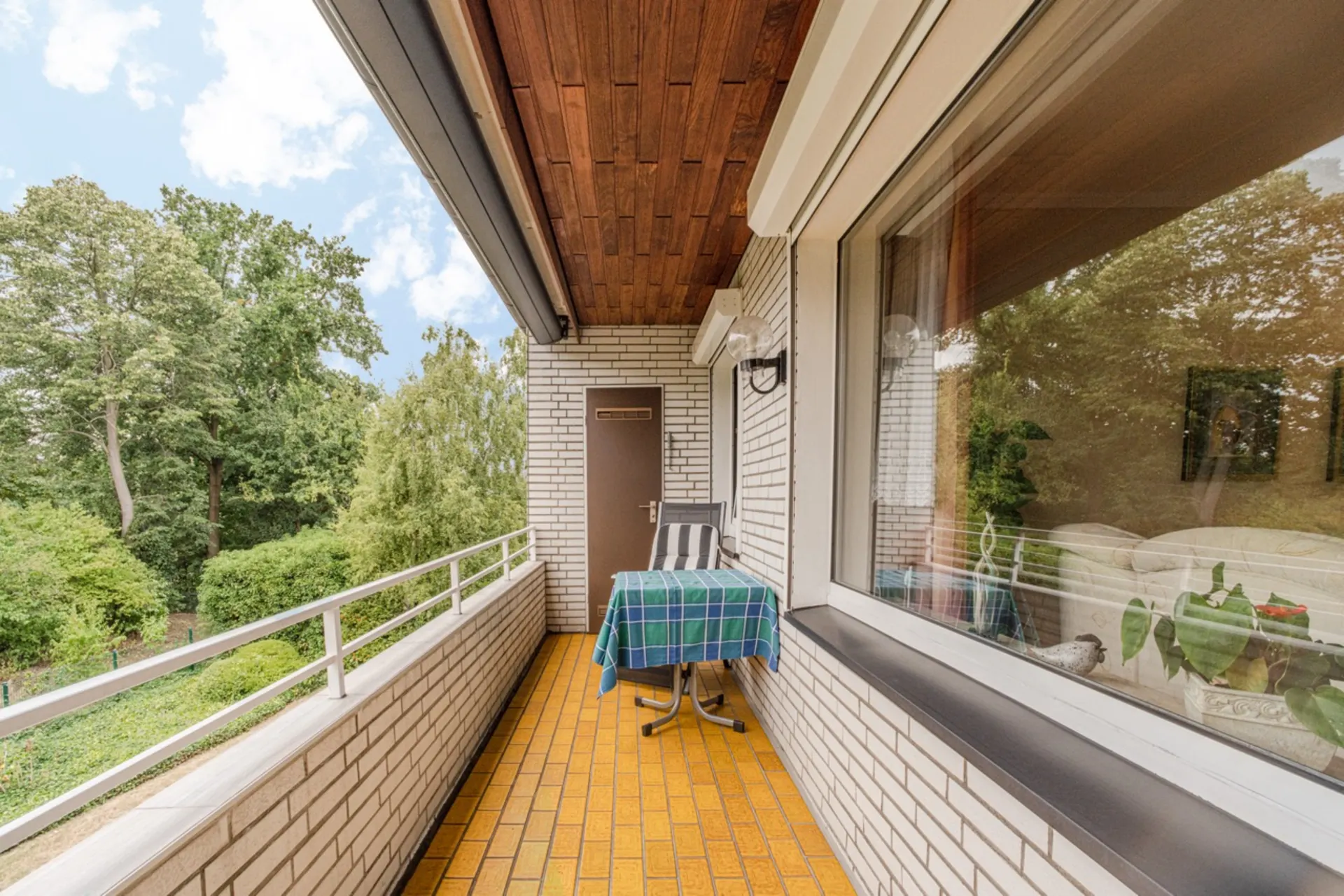 Balkon mit gelben Bodenfliesen, Tisch mit blau-grünem Tuch, Stuhl und Blick ins Grüne.