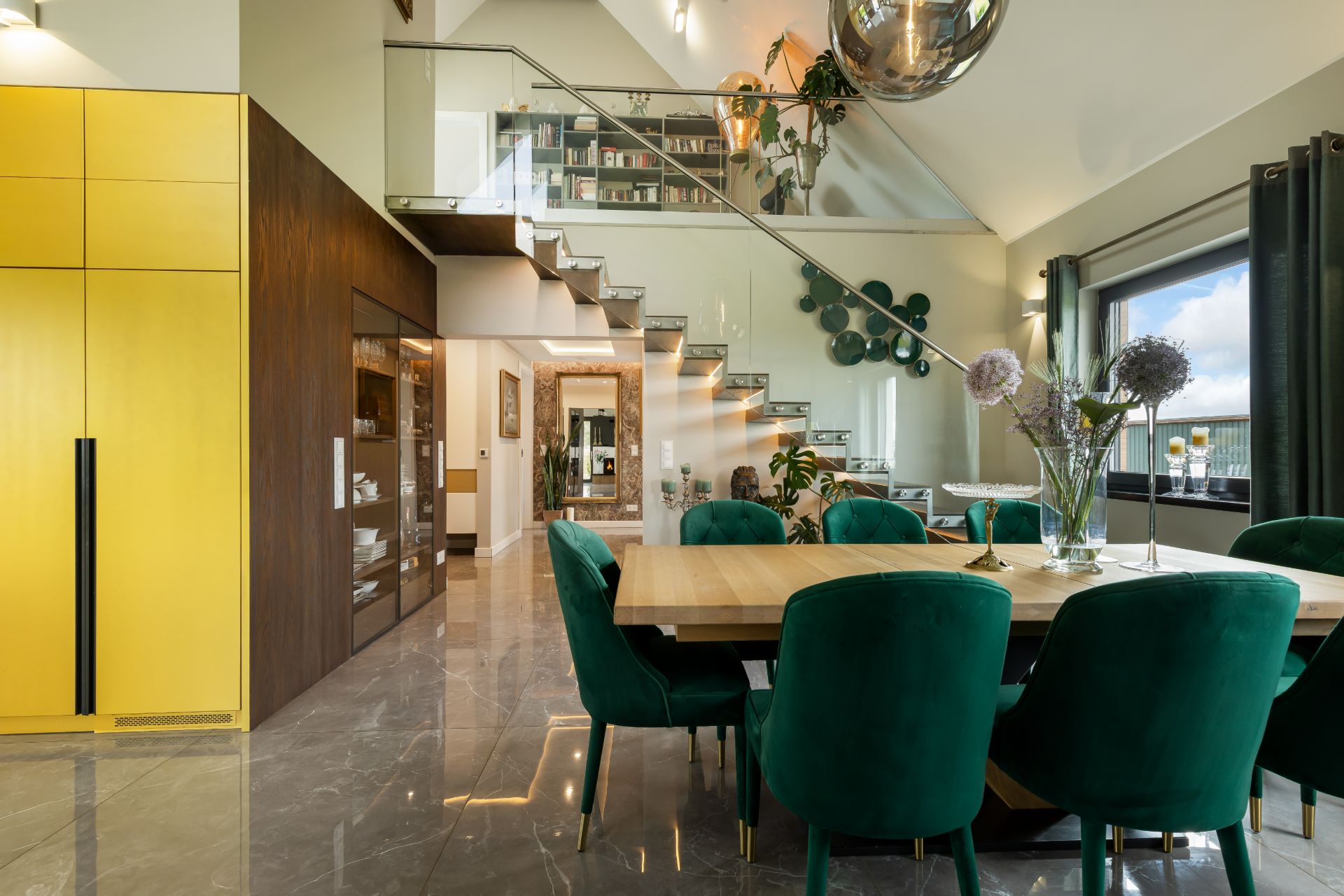 Modernes Esszimmer mit grünem Stuhlset, Holztisch und gelben Schränken, offene Treppe und Glasgeländer.
