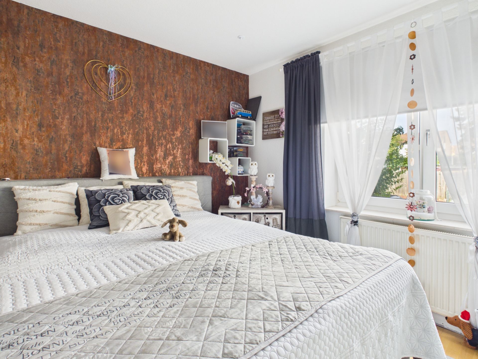 Helles Schlafzimmer mit großem Bett, Kissen, brauner Akzentwand und Fenster mit weißen Vorhängen.