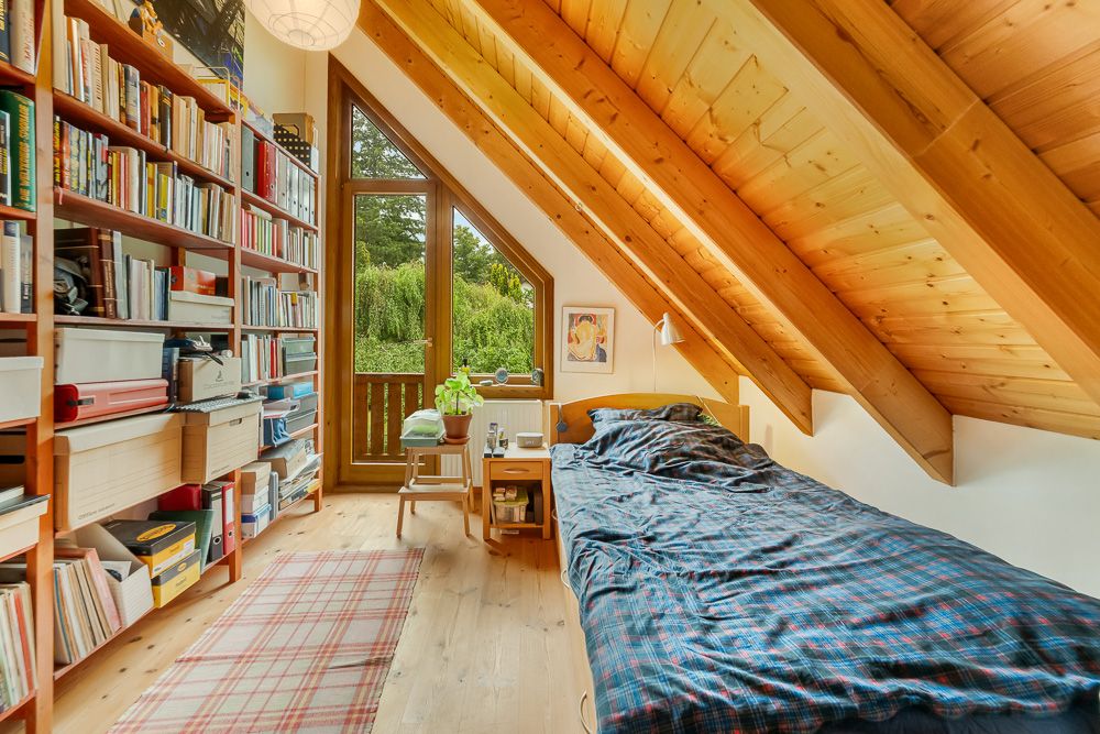 Gemütliches Dachzimmer mit Einzelbett, Bücherregal, Holzboden und großem Fenster mit Blick ins Grüne.