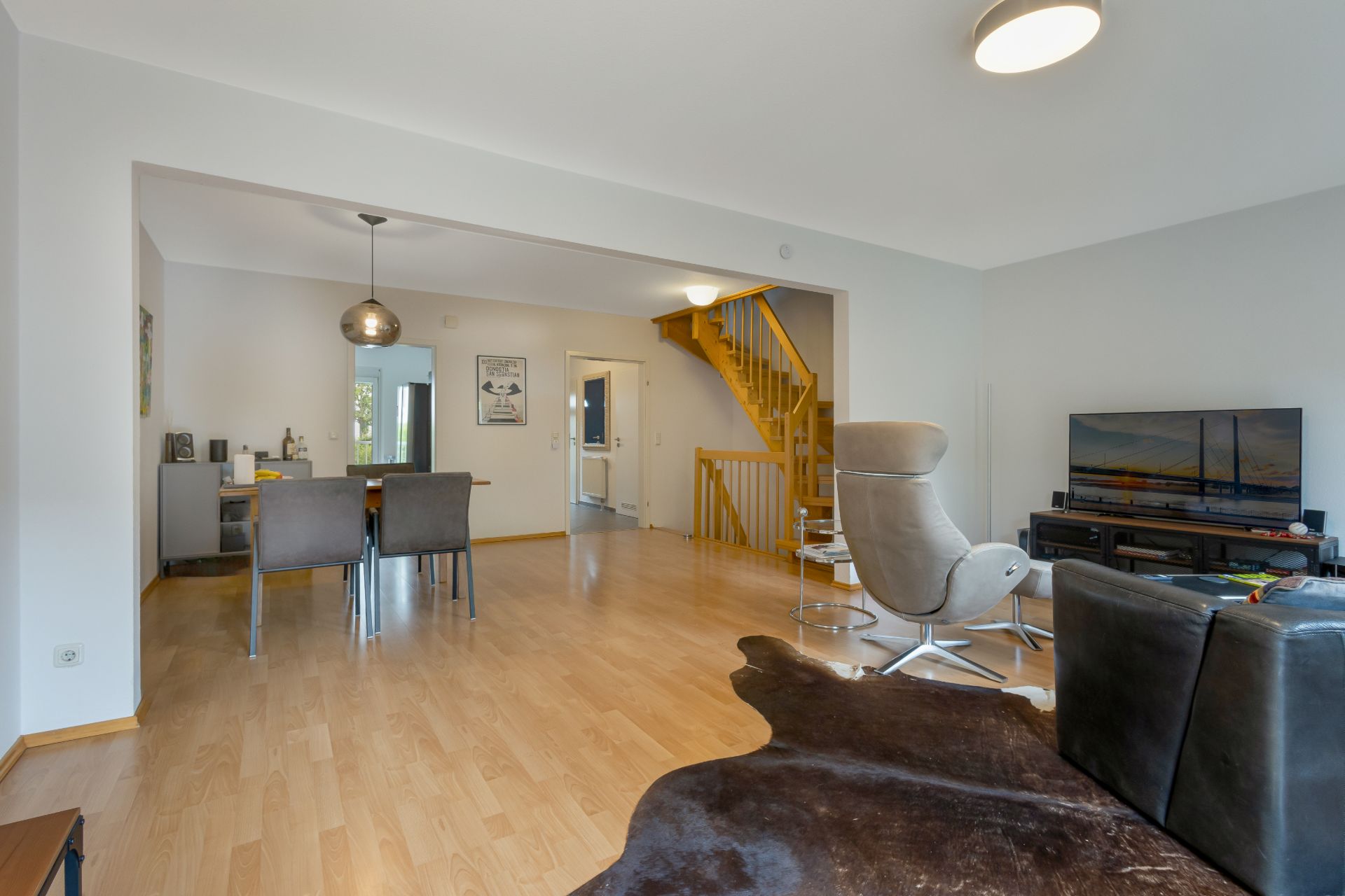Helles Wohnzimmer mit Holzboden, Essbereich, TV, modernen Möbeln und einer Treppe im Hintergrund.