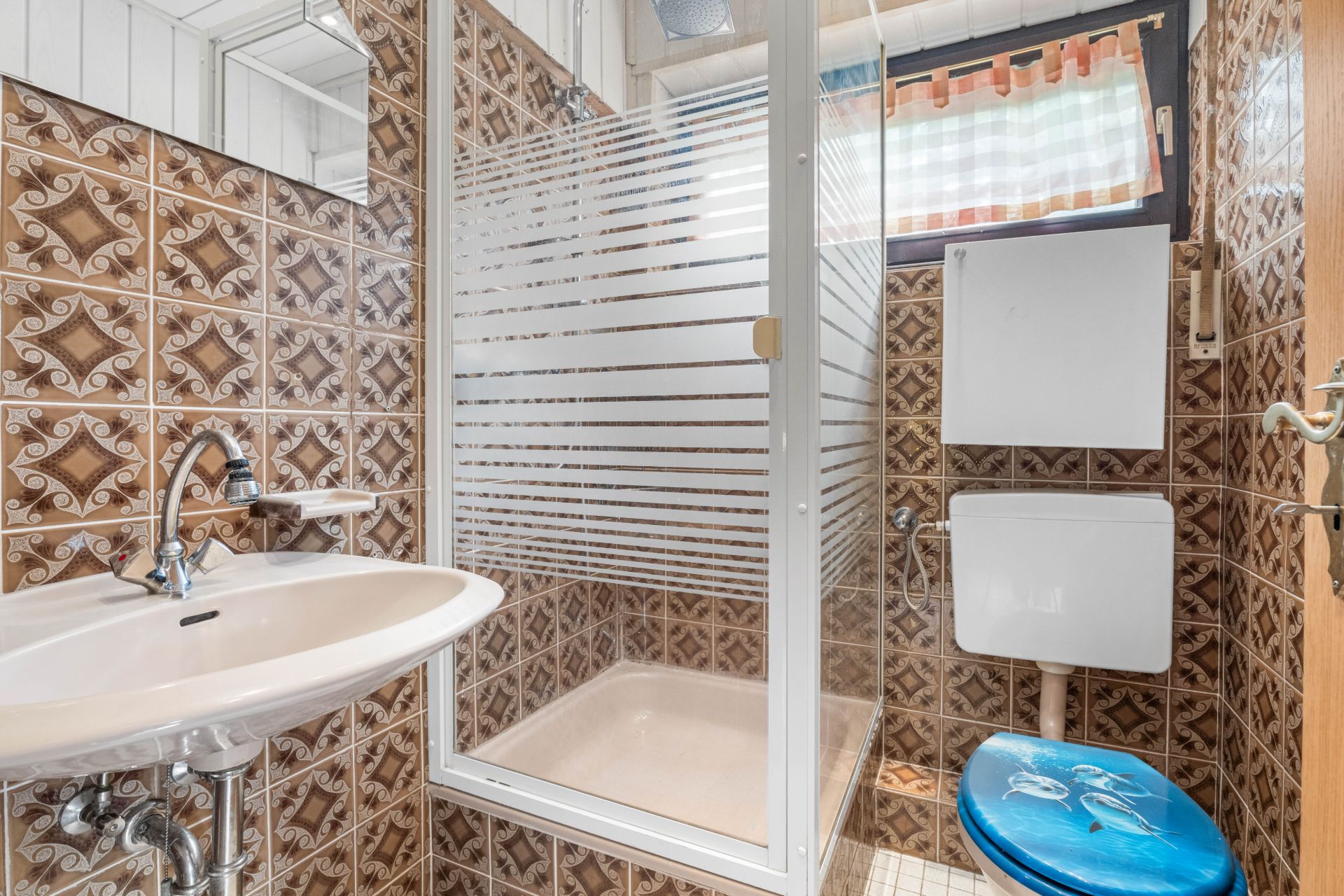 Kleines Bad mit gemusterter Fliesenwand, Dusche, Waschbecken und Toilette mit blauem Sitz.