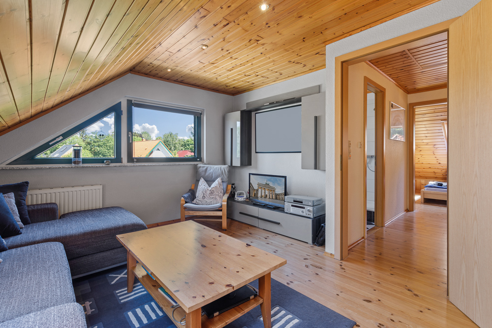 Wohnzimmer mit Holzdecke, großem Fenster, Sofa, Couchtisch und TV-Möbel, Zugang zum Flur.