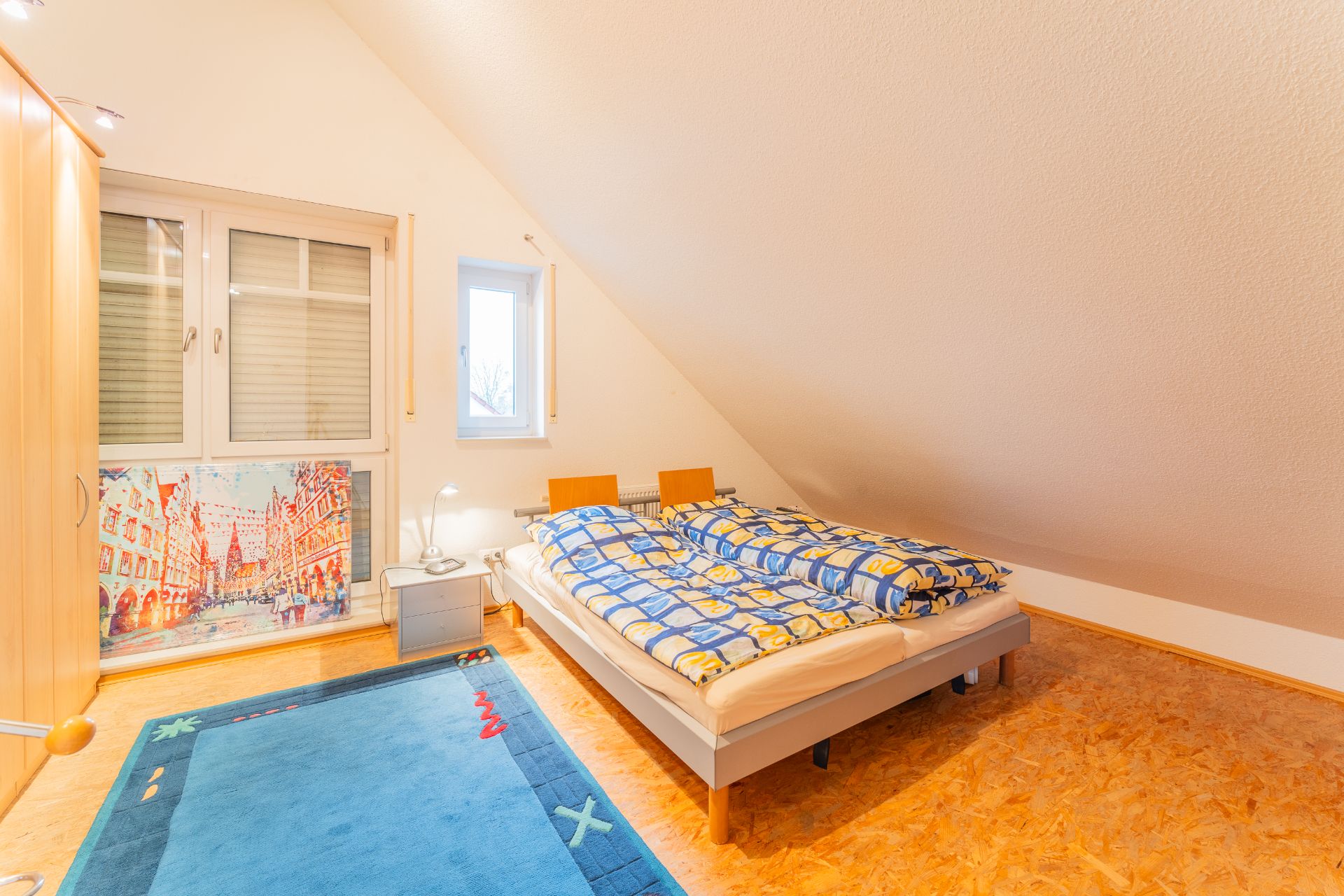 Helles Schlafzimmer mit Doppelbett, blauem Teppich, Schrank und Fenster unter einer Dachschräge.