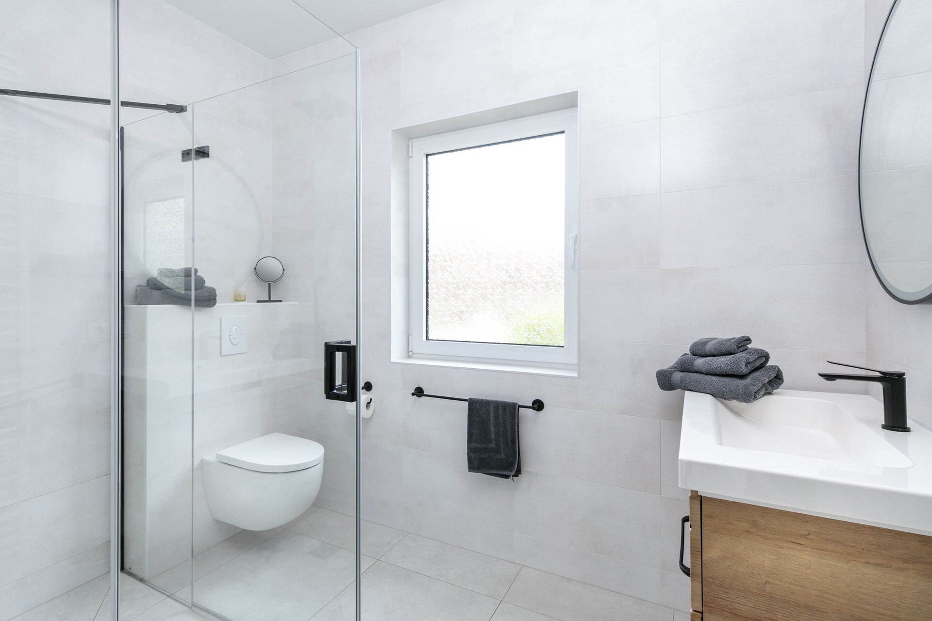 Modernes Badezimmer mit Glasdusche, WC, Fenster, Waschbecken und Handtüchern auf der Ablage.