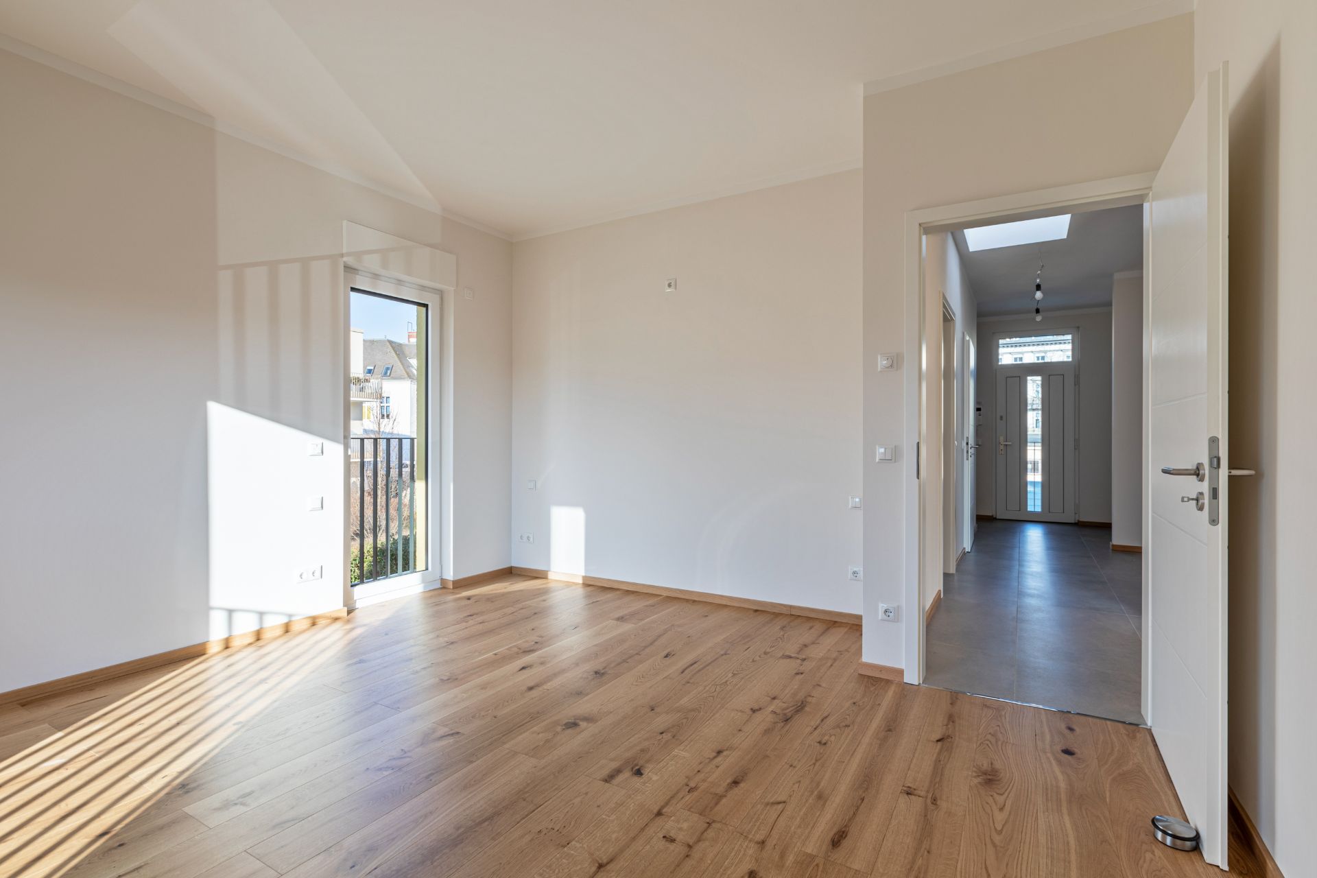 Helles, leeres Zimmer mit Holzfußboden und bodentiefem Fenster, Eingangstür im Hintergrund.