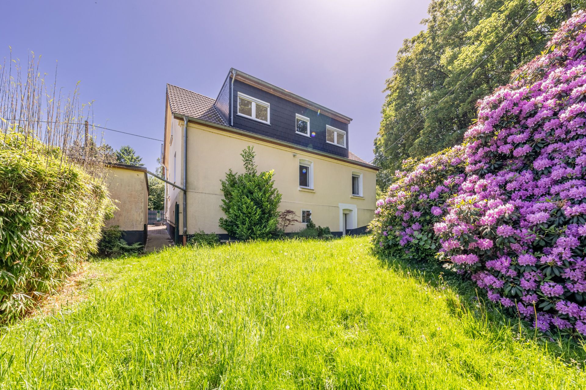 Gelbes Wohnhaus mit dunklem Dach, grünem Garten und blühenden Sträuchern bei sonnigem Wetter.