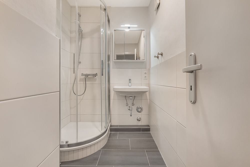 Modernes Badezimmer mit Dusche, Waschbecken und Spiegel, graue Fliesen, helle Wände.