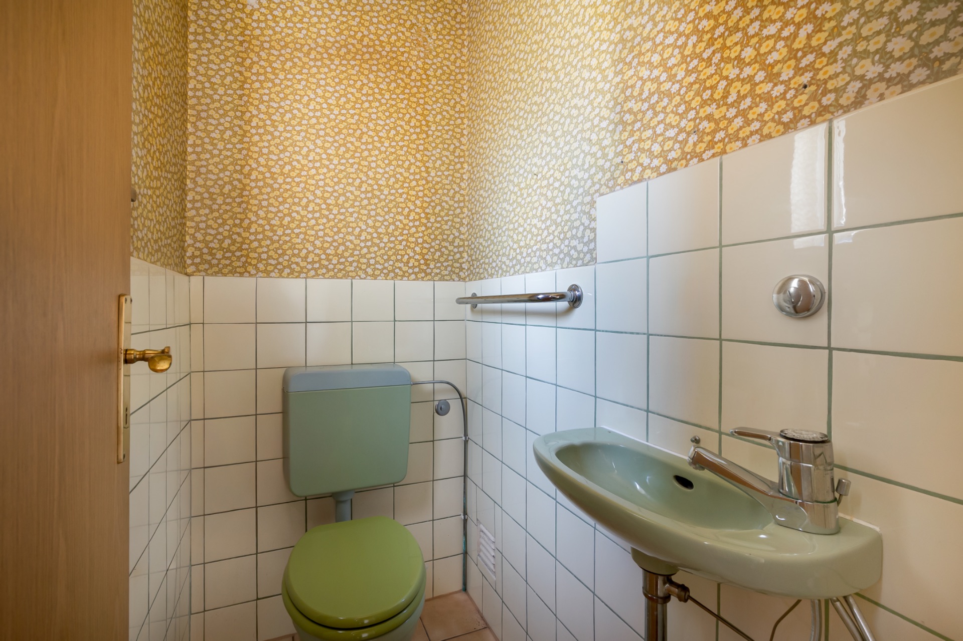 Kleines WC mit grünem WC, Waschbecken und gemusterter Tapete an den Wänden.