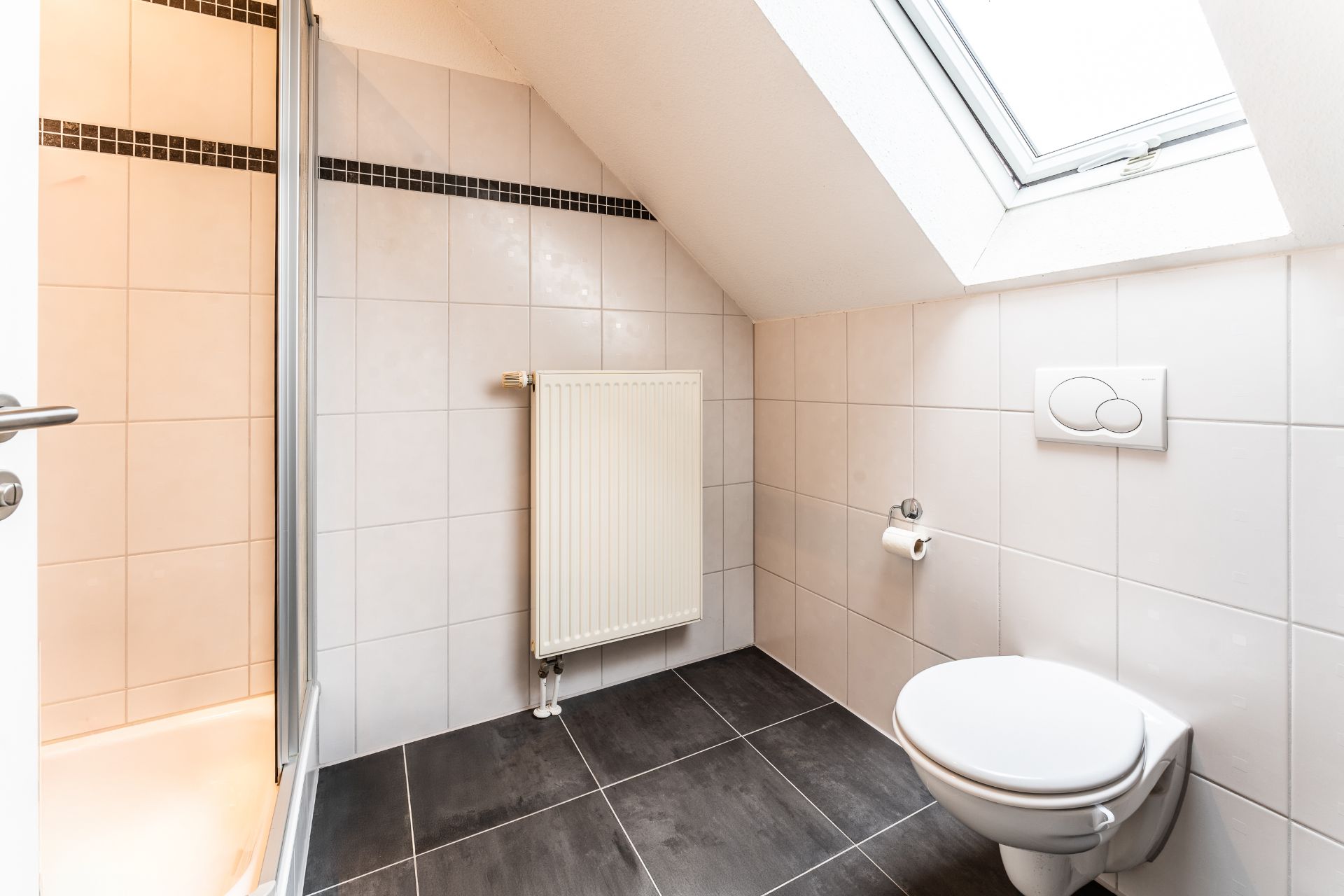 Helles Badezimmer mit Dusche, Wandtoilette, Heizkörper und Dachfenster.