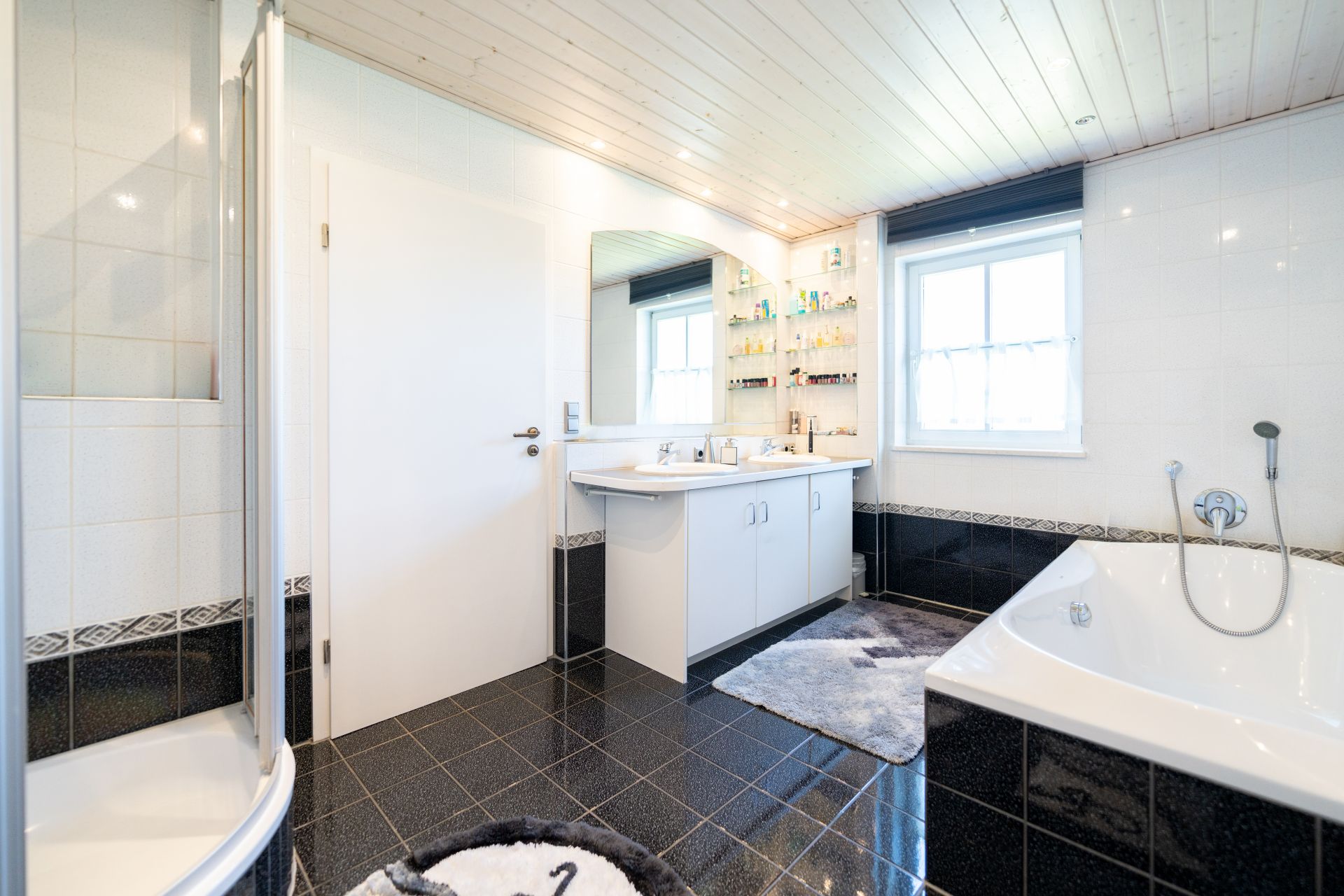 Modernes Badezimmer mit Badewanne, Dusche, Doppelwaschbecken, Fenster und dunklem Fliesenboden.