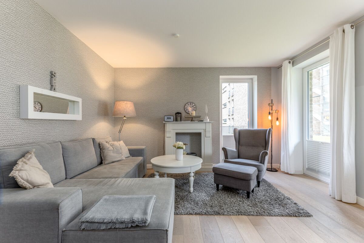 Helles Wohnzimmer mit grauem Sofa, Sessel, Kamin, Couchtisch und großen Fenstern.