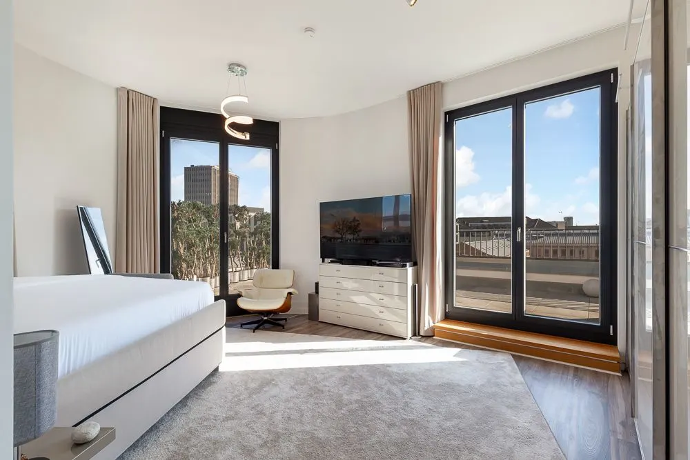 Helles Schlafzimmer mit großem Bett, TV, Sessel und bodentiefen Fenstern zu einem Balkon.