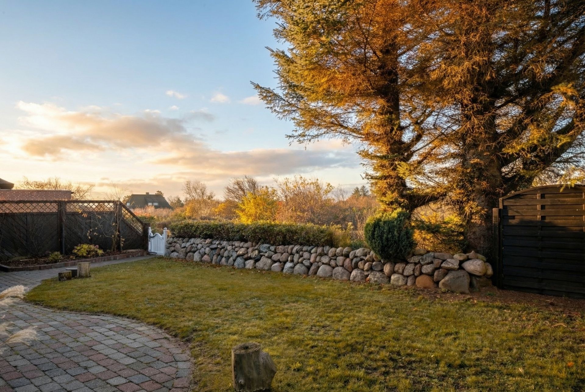 Garten mit Rasenfläche, Pflasterweg, Steinmauer und großen Bäumen bei Sonnenuntergang.