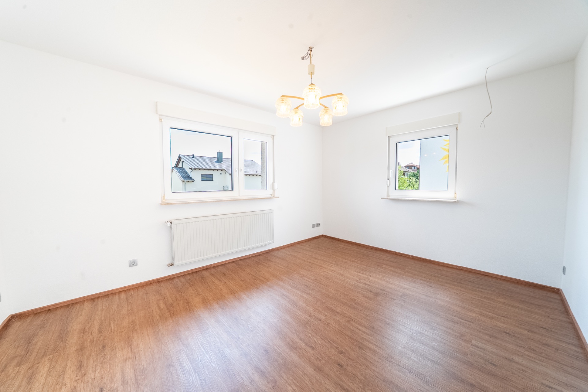 Helles, leeres Zimmer mit Holzfußboden, zwei Fenstern und Deckenleuchte.