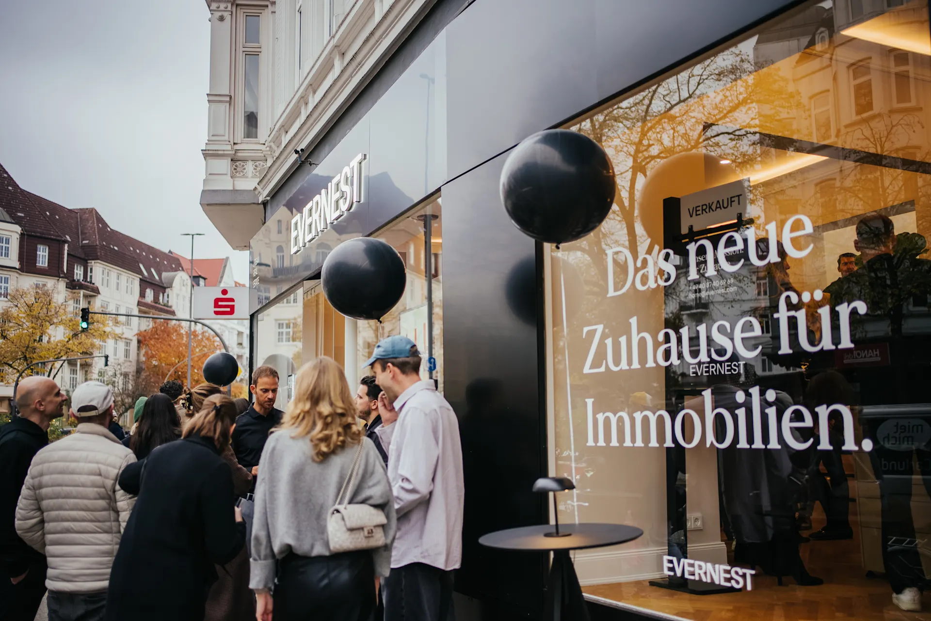 Menschen stehen vor einem Geschäft mit großer Fensterbeschriftung und schwarzen Ballons.