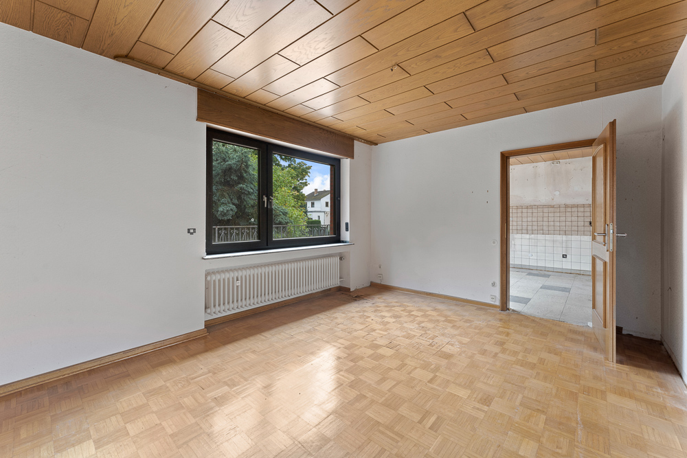 Leerer Raum mit Parkettboden, Holzdecke, großem Fenster und geöffneter Tür zur gefliesten Küche.