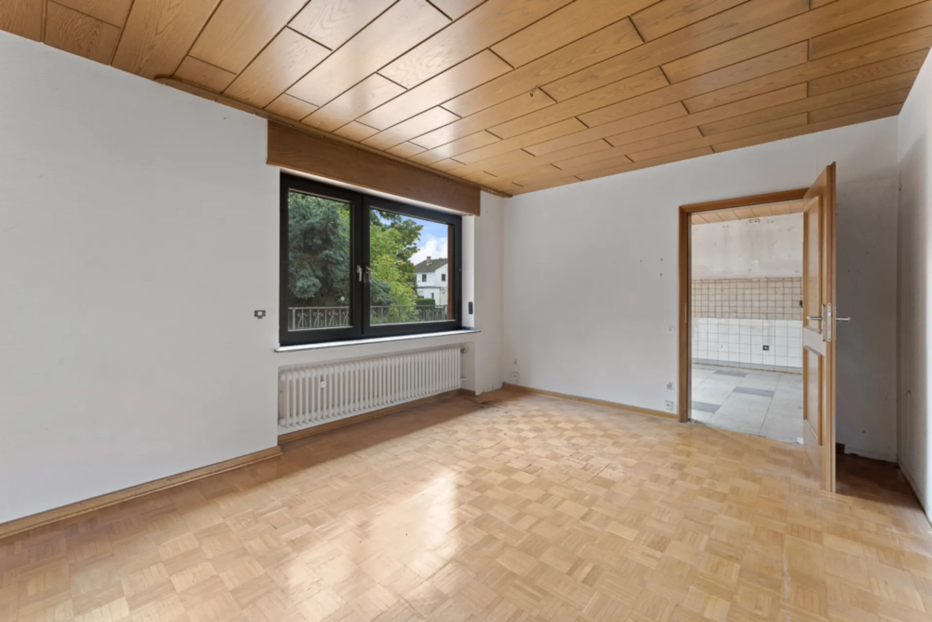 Leerer Raum mit Parkettboden, Holzdecke, großem Fenster und geöffneter Tür zur gefliesten Küche.