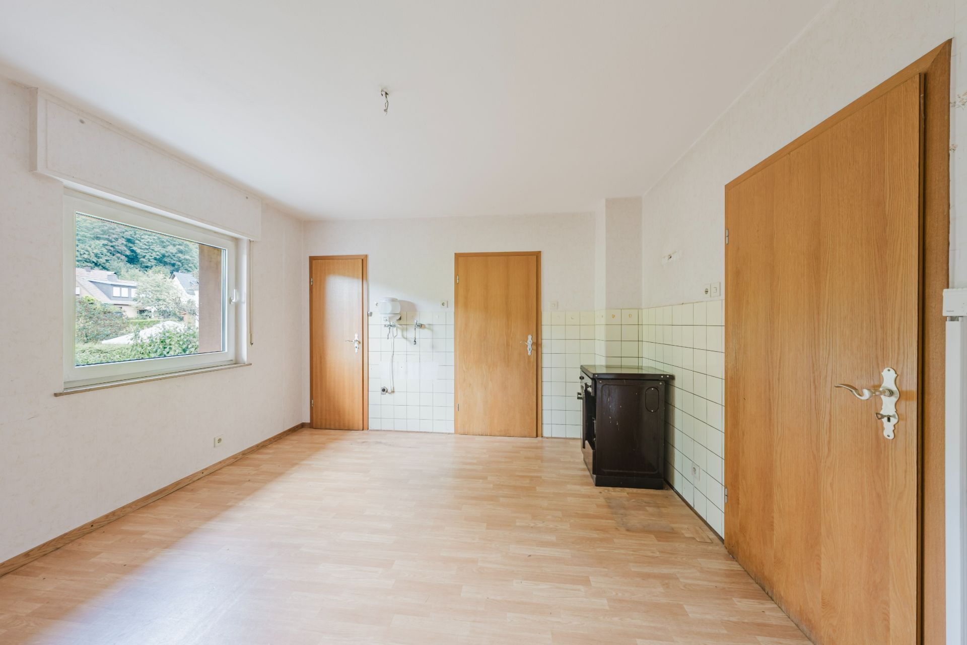 Leerer Raum mit Holzfußboden, drei Holztüren, großem Fenster und kleiner Küchenecke.