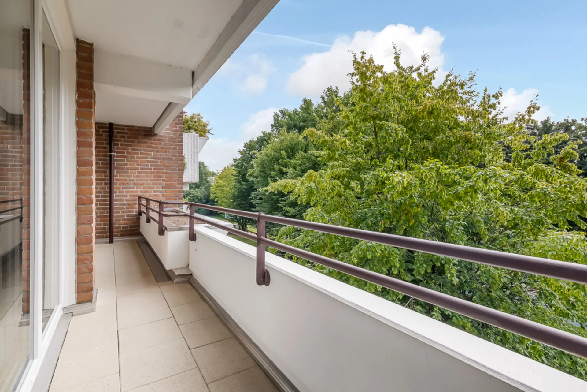 Langer Balkon mit Fliesenboden, Metallgeländer und Blick auf grüne Bäume.
