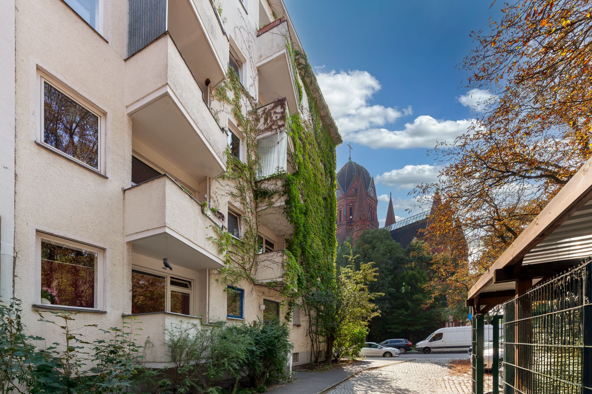 Wohnhaus mit Balkonen und begrünten Fassaden, davor Bäume und geparkte Autos.