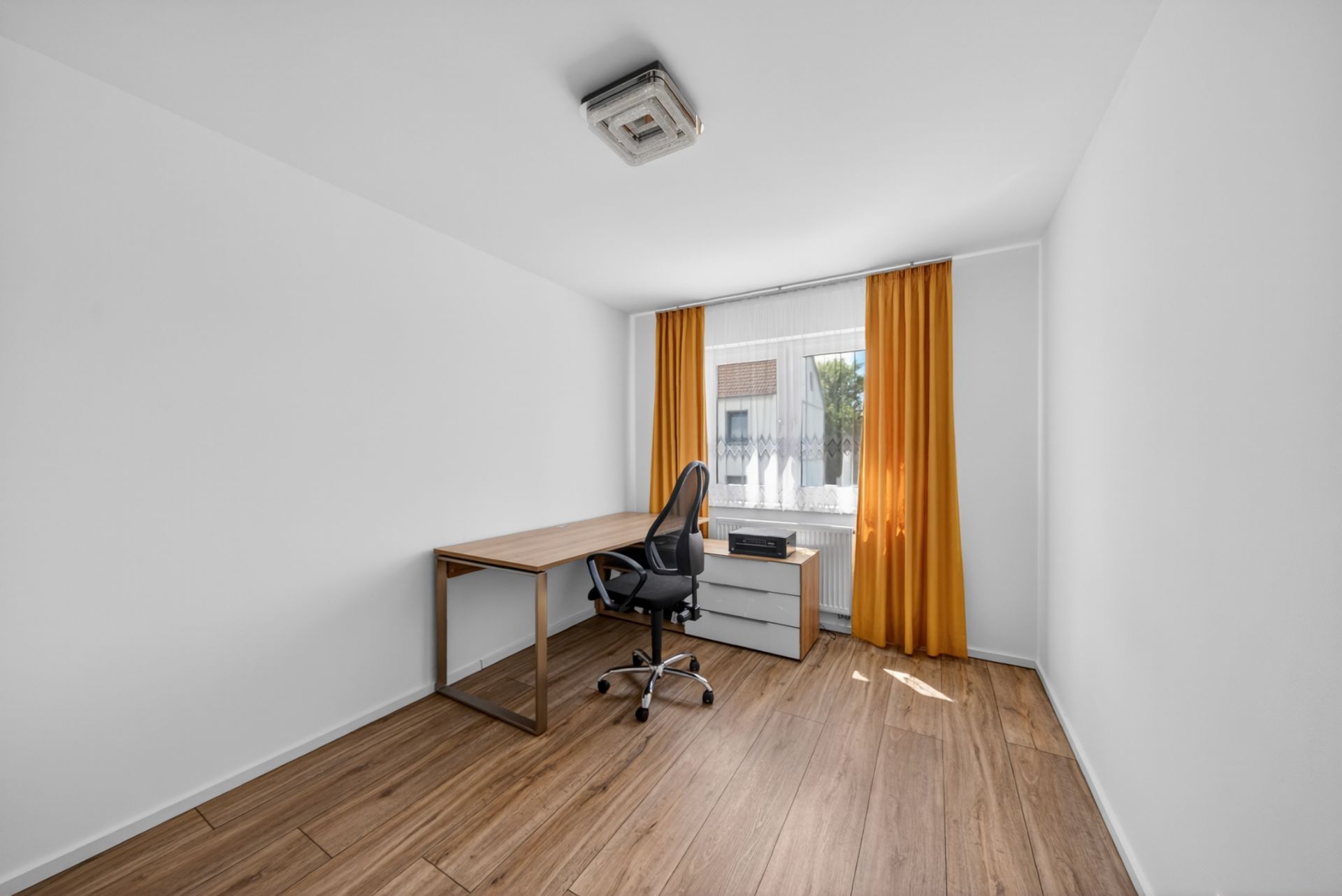 Helles Arbeitszimmer mit Schreibtisch, Stuhl, Kommode und gelben Vorhängen vor dem Fenster.
