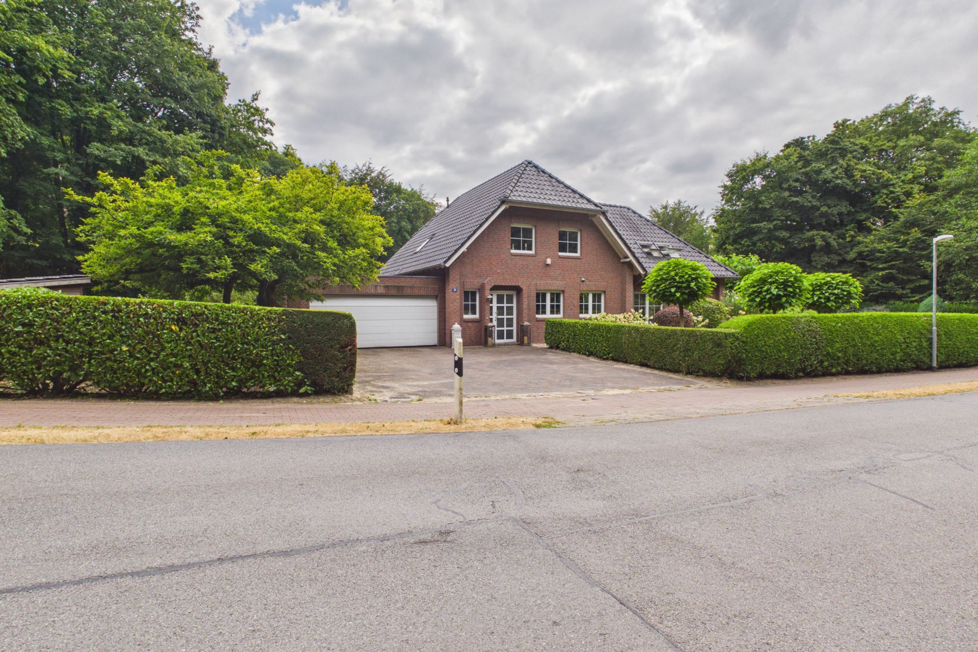 Backsteinwohnhaus mit Spitzdach, Garage und gepflegtem Garten, von Hecken umgeben.