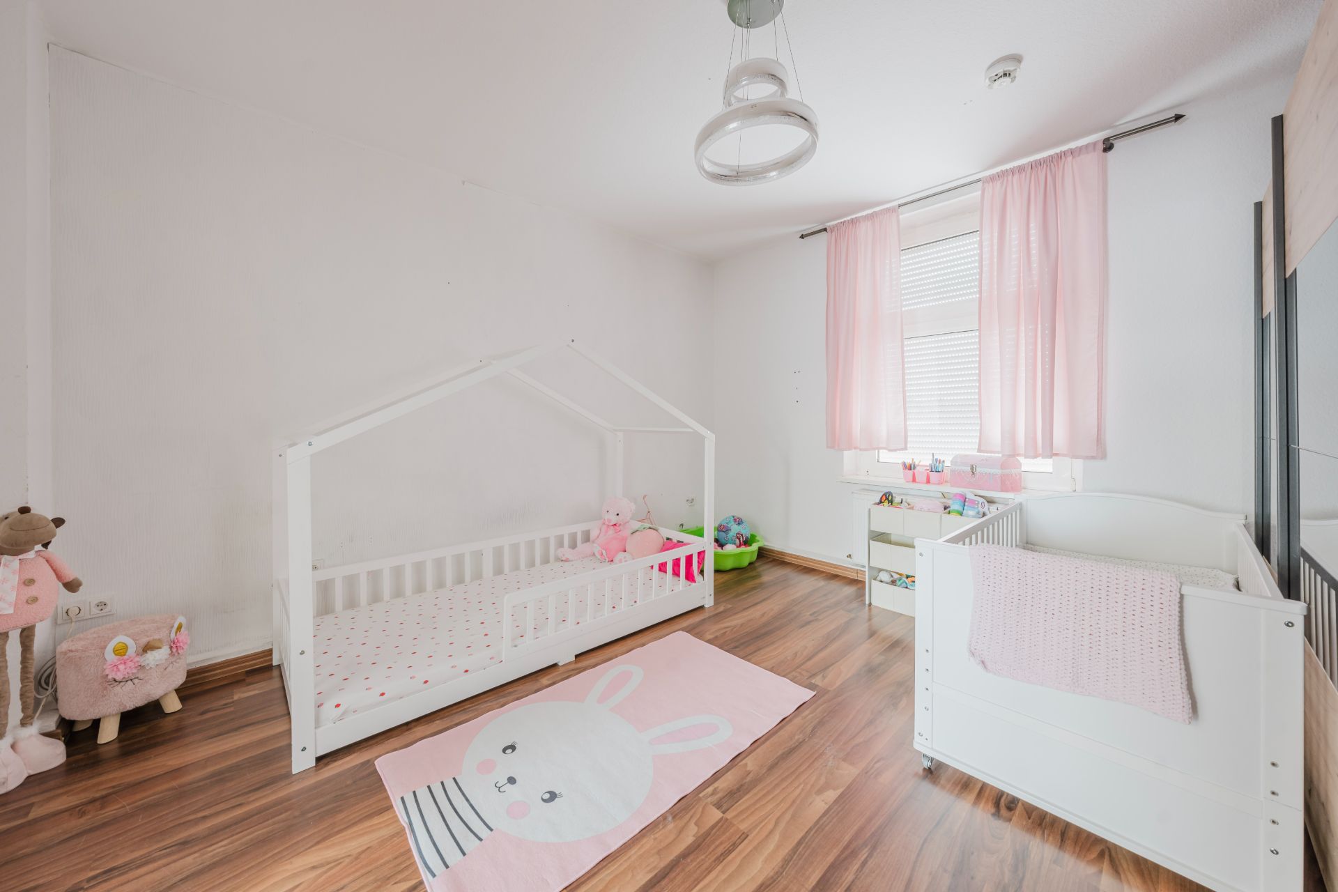 Helles Kinderzimmer mit Holzboden, Gitterbett, Kinderbett und rosa Vorhängen.