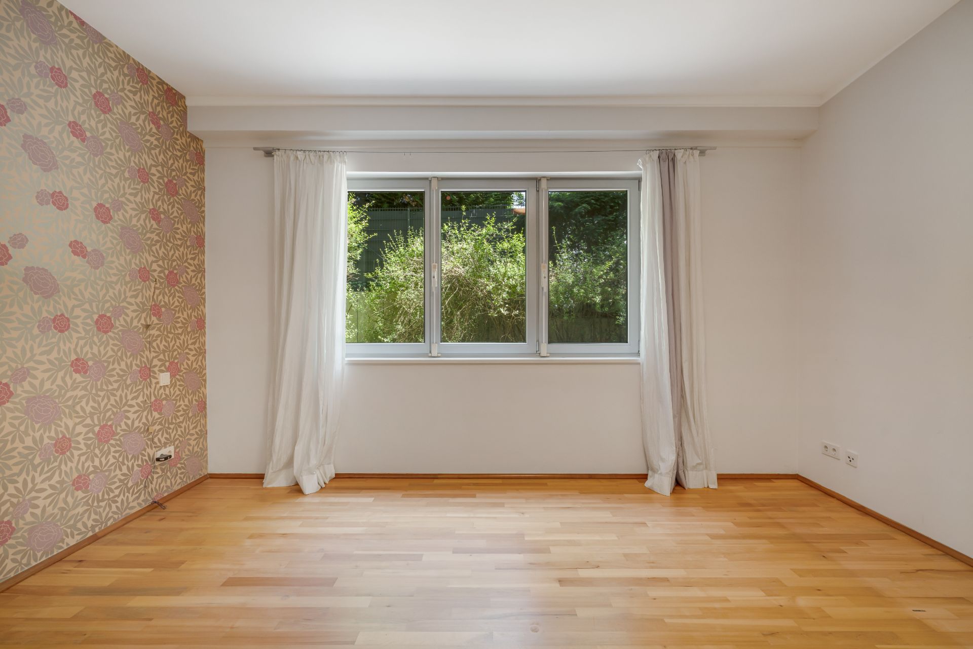 Leerer Raum mit Holzfußboden, Fenster mit weißen Vorhängen und gemusterter Tapete an einer Wand.