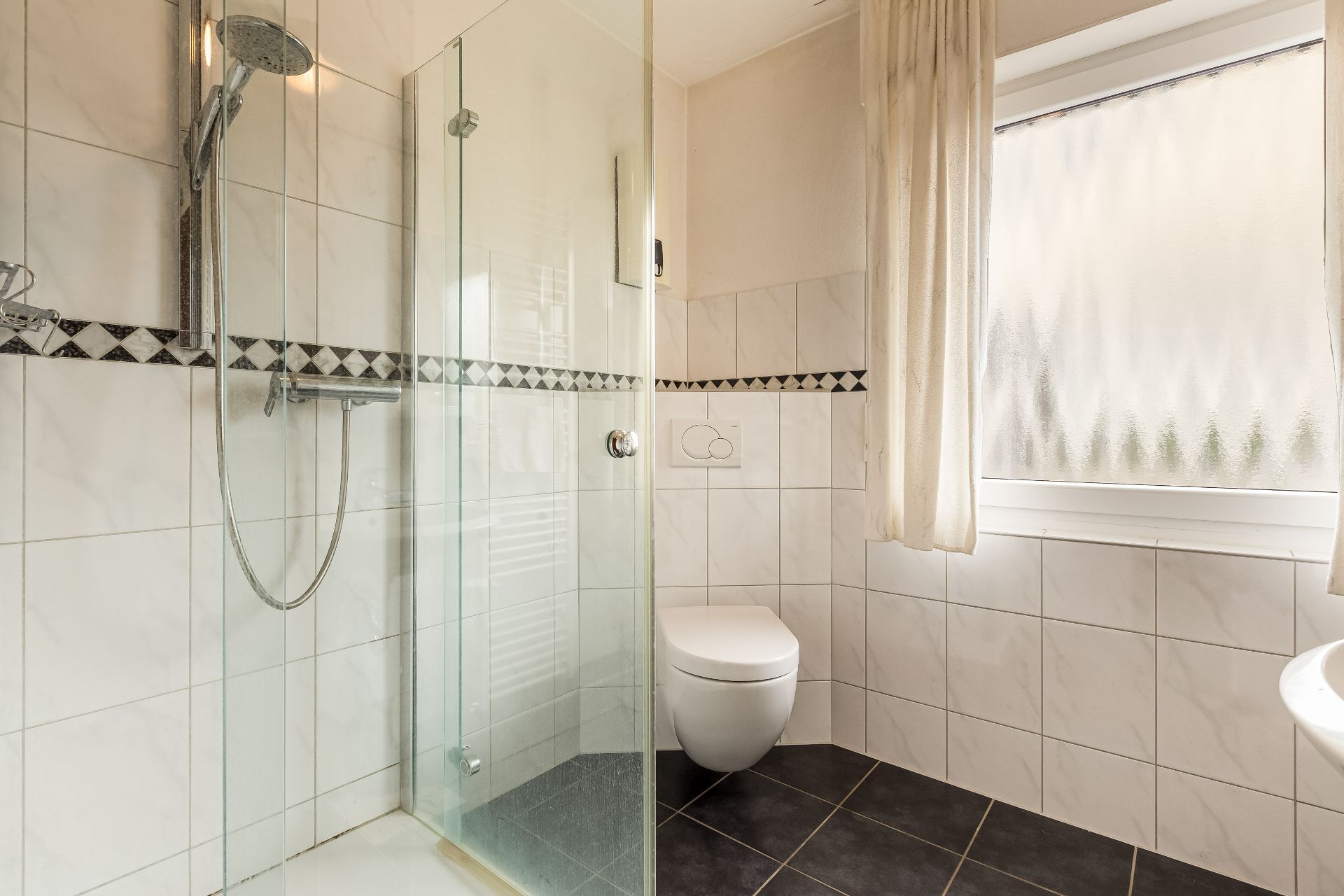 Modernes Bad mit ebenerdiger Dusche, WC und großem Fenster mit Vorhang.