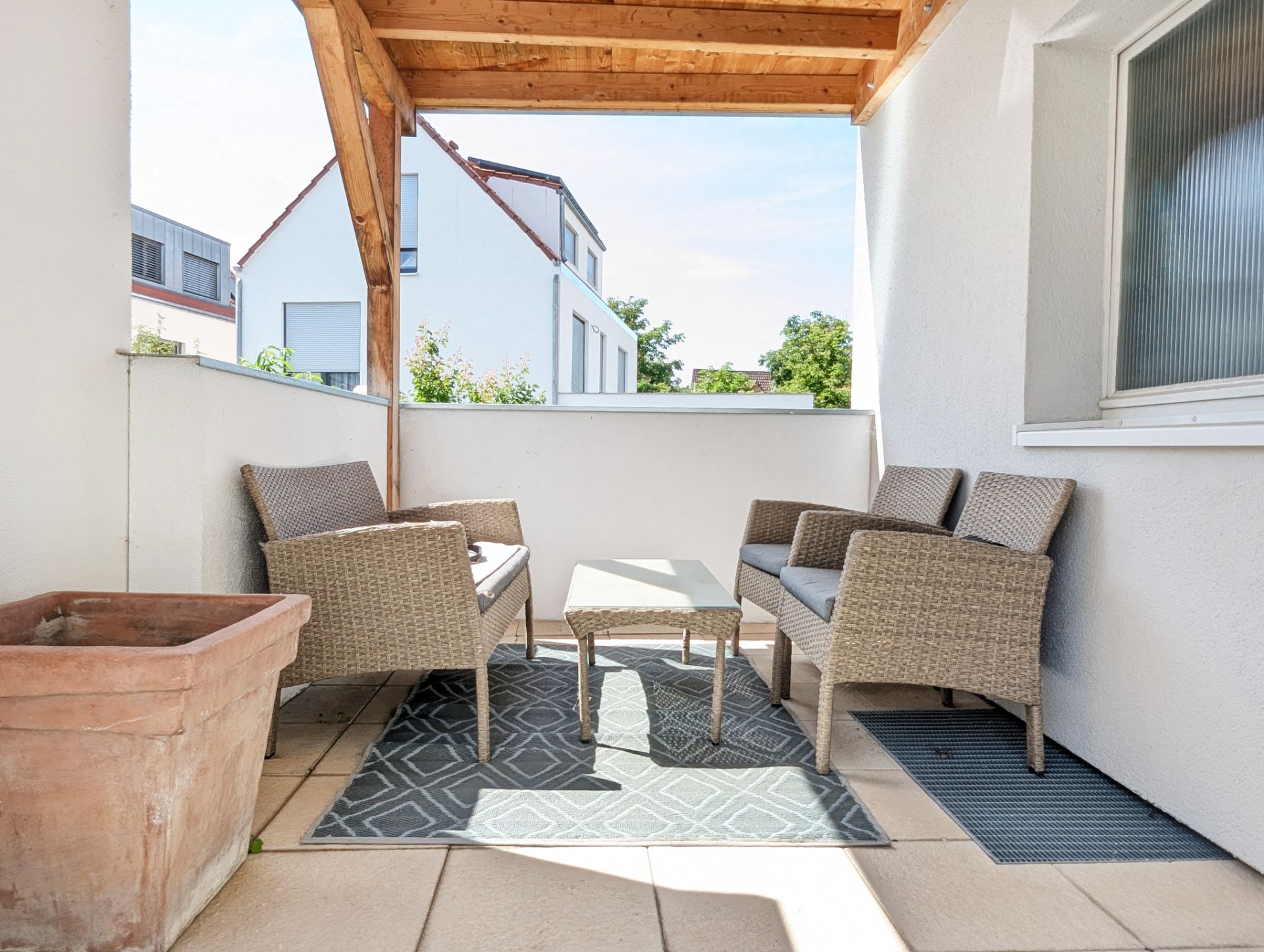 Überdachter Balkon mit zwei Rattansofas, Tisch, Teppichen und großem Blumenkübel.