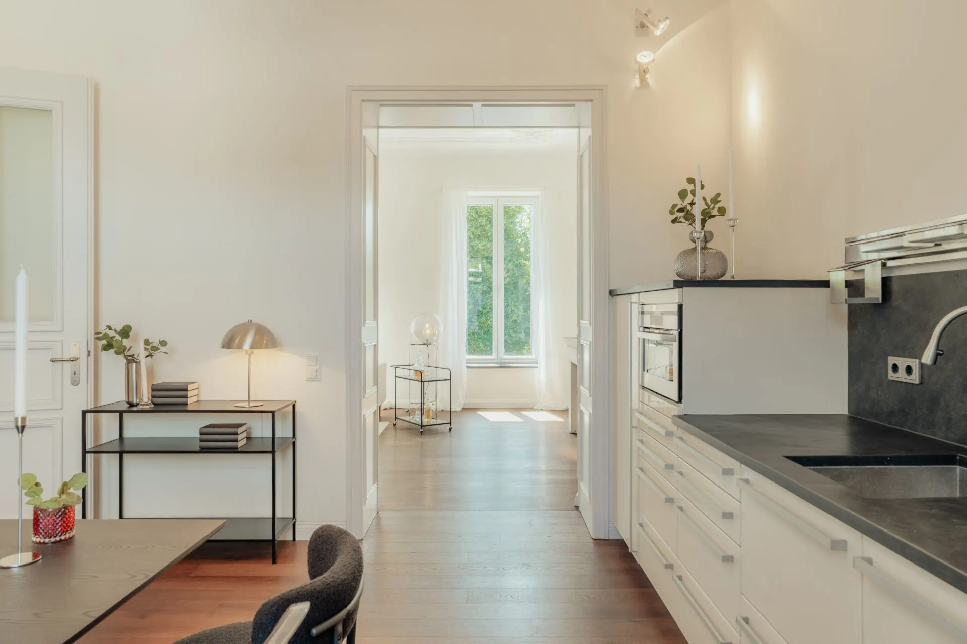 Helle Küche mit weißen Schränken, Tisch und Blick in ein weiteres Zimmer mit Fenster.