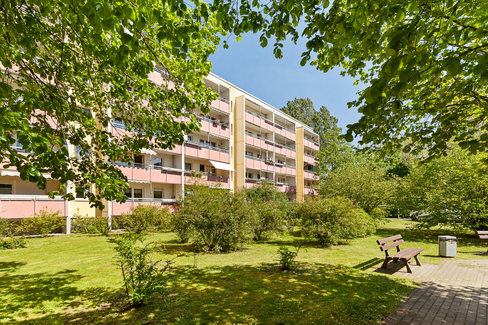 Mehrfamilienhaus mit Balkonen, gepflegter Garten und Bänken im Vordergrund bei sonnigem Wetter.