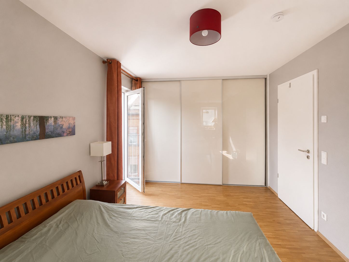 Helles Schlafzimmer mit Holzfußboden, Doppelbett, Kleiderschrank und offenem Fenster.