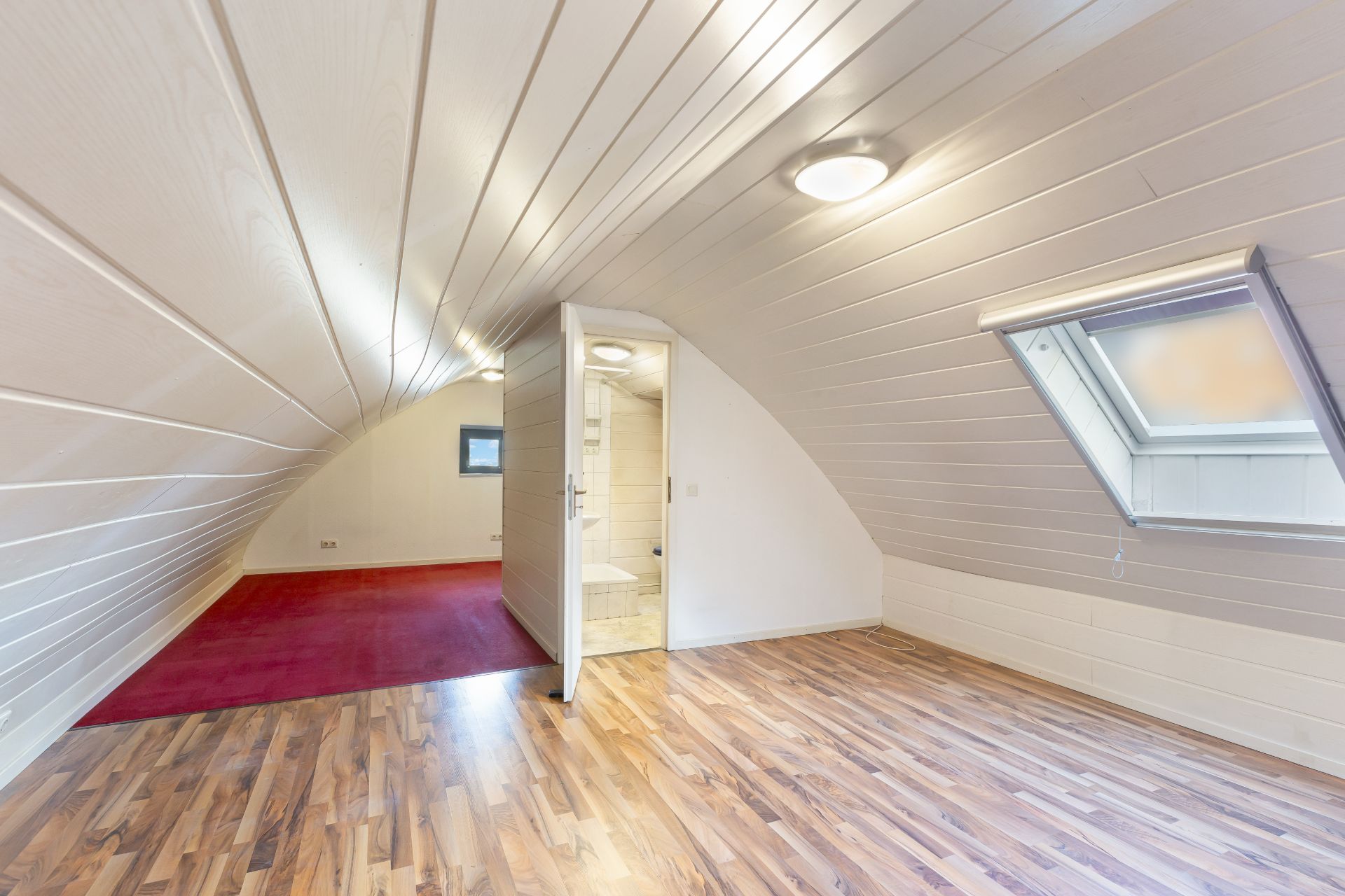 Dachzimmer mit Holzfußboden, rotem Teppichbereich, Fenster und offenem Bad mit Toilette.